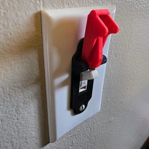 May include: A red light switch extender attached to a black switch plate. The switch plate is mounted on a white wall. The switch is in the "ON" position.