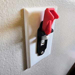 May include: A red and black light switch cover installed on a white wall plate. The switch is in the "ON" position. The cover is designed to prevent accidental activation of the switch.