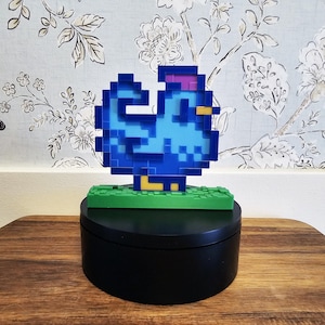 May include: A pixelated blue rooster figurine with a purple comb and yellow beak, standing on a green base. The figurine is displayed on a black rotating stand, set against a floral wallpaper background.