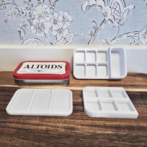 May include: A rectangular metal Altoids peppermint tin with a red and white lid, accompanied by three white plastic paint palette inserts. Two of the inserts feature six rectangular wells, while the third insert contains three long rectangular wells.