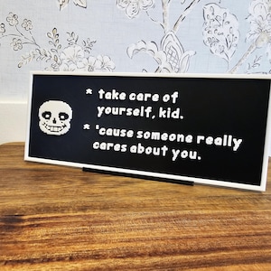 May include: A black sign with white text that reads "take care of yourself, kid. 'cause someone really cares about you." A white pixelated skull is on the left. The sign is in a white frame and sits on a wooden surface.