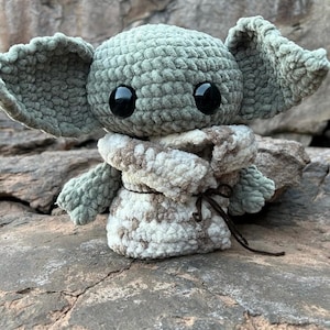 Puede incluir: Un peluche de ganchillo que se asemeja a un personaje con orejas grandes y ojos negros. El juguete es principalmente verde claro con una envoltura estampada en blanco y marr&oacute;n. El juguete est&aacute; sobre una superficie rocosa gris texturizada.