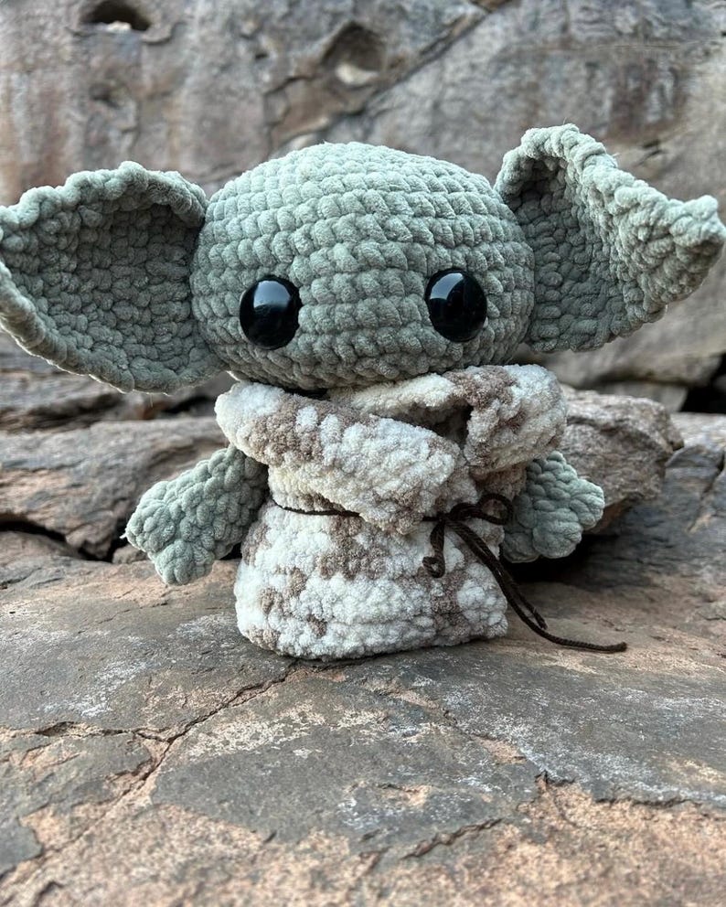 Puede incluir: Un peluche de ganchillo que se asemeja a un personaje con orejas grandes y ojos negros. El juguete es principalmente verde claro con una envoltura estampada en blanco y marr&oacute;n. El juguete est&aacute; sobre una superficie rocosa gris texturizada.