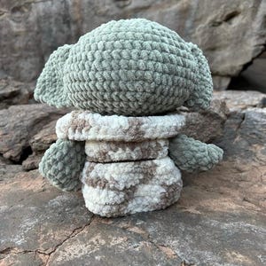 Puede incluir: Mu&ntilde;eco de ganchillo hecho a mano, parecido a un personaje con orejas grandes. El mu&ntilde;eco tiene cabeza y orejas de color verde claro, y un cuerpo con estampado blanco y marr&oacute;n. El mu&ntilde;eco est&aacute; sobre una roca.
