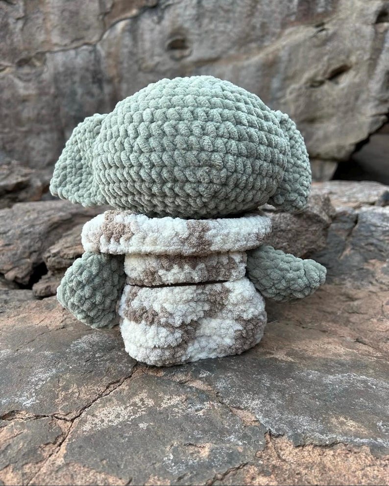 Puede incluir: Mu&ntilde;eco de ganchillo hecho a mano, parecido a un personaje con orejas grandes. El mu&ntilde;eco tiene cabeza y orejas de color verde claro, y un cuerpo con estampado blanco y marr&oacute;n. El mu&ntilde;eco est&aacute; sobre una roca.
