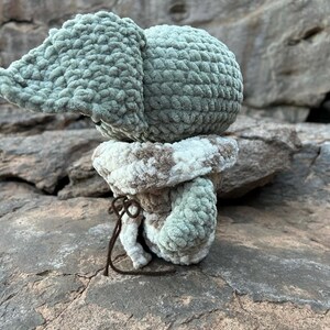 Puede incluir: Un elefante de ganchillo hecho a mano en tonos verdes, blancos y marrones. El elefante tiene orejas grandes y un cuerpo redondo. La base del elefante es un patr&oacute;n a rayas blanco y marr&oacute;n. Una cuerda marr&oacute;n est&aacute; atada alrededor de la base.