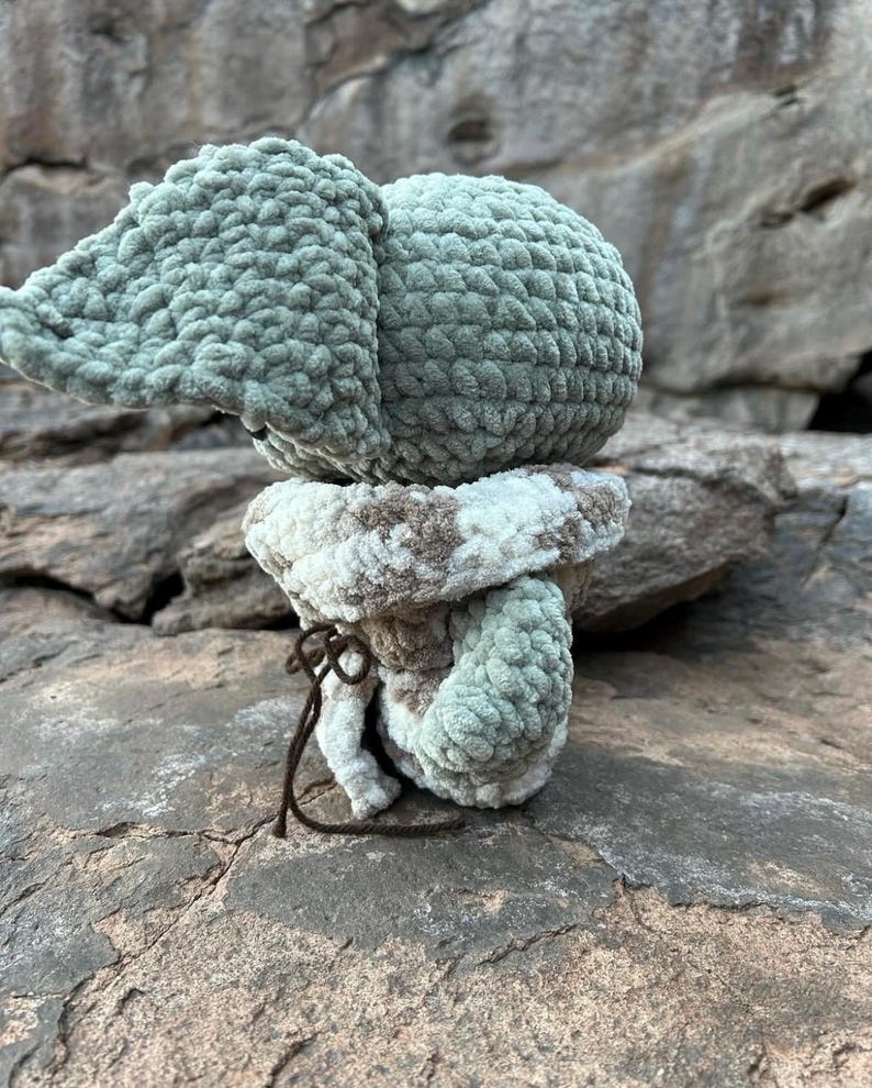 Puede incluir: Un elefante de ganchillo hecho a mano en tonos verdes, blancos y marrones. El elefante tiene orejas grandes y un cuerpo redondo. La base del elefante es un patr&oacute;n a rayas blanco y marr&oacute;n. Una cuerda marr&oacute;n est&aacute; atada alrededor de la base.