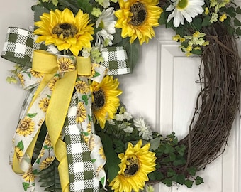 May include: A green and white checkered ribbon with a yellow ribbon tied into a bow, decorated with sunflowers and greenery. The wreath is made of brown vines.