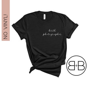 May include: Black t-shirt with the text "birth photographer" in white lettering. The shirt is tied in a knot at the bottom. The image also includes a logo with the letters "B+B" in a circle.