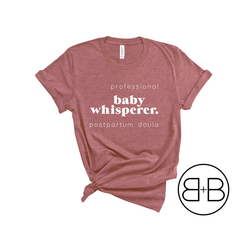 May include: A dusty rose-colored t-shirt with the words "professional baby whisperer. postpartum doula" in white. The shirt is knotted at the bottom. A circular logo with the letters "B + B" is in the lower right corner.