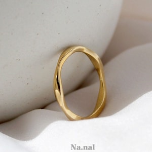 May include: A gold-toned, twisted, ring with a smooth, rounded, oval shape. The ring is on a white fabric background.