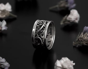 Puede incluir: Un anillo de plata con una banda ancha que presenta un diseño intrincado y ornamentado de hojas y vides en espiral.