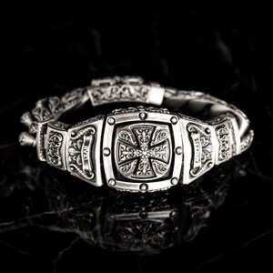 May include: A silver bracelet with a detailed cross design in the center. The bracelet is made of multiple links with intricate floral patterns.