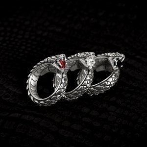Könnte beinhalten: Drei silberne Schlangenringe mit verschiedenen Edelsteinen. Der Ring links hat einen roten Edelstein, der mittlere Ring einen weißen Edelstein und der Ring rechts einen schwarzen Edelstein.