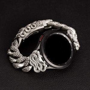 Könnte beinhalten: Ein silberfarbenes Smartwatch-Armband mit detailliertem Schlangendesign. Das Armband umschließt eine schwarze Smartwatch und zeigt die komplizierten Schuppen und Schlangenkopfdetails. Das Zifferblatt der Uhr ist schwarz, und das Armband scheint aus Metall zu sein.