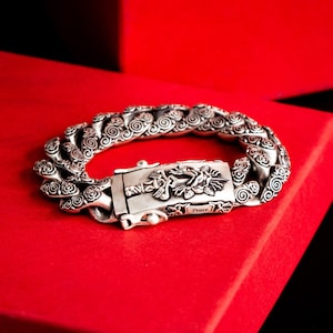May include: A silver bracelet with a detailed design of swirling patterns and roses. The clasp features a sword and rose design with the word "Peace" inscribed on it.
