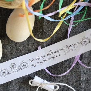 May include: A white paper strip with black calligraphy that reads "Ask and you shall receive, that your joy may be complete. John 16.23". The strip is decorated with a black floral design. Two white wooden eggs and colorful ribbons are in the background.