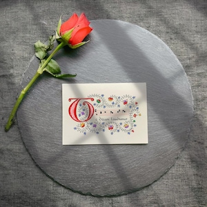 Puede incluir: Una tarjeta rectangular con una gran letra O roja iluminada, notación musical en un pentagrama rojo y el texto Te Deum laudamus, rodeada de coloridas enredaderas florales, colocada junto a una rosa naranja sobre una pizarra redonda.