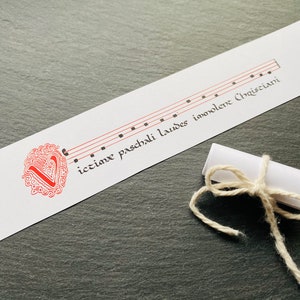 May include: A white card with a red and white decorative design and the text "ictuine paschali laudes inmoventer Christiani" in black calligraphy. The card has a musical staff with notes on it.