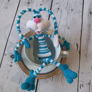 Puede incluir: Un conejo blanco de crochet con brazos y piernas a rayas azules y blancas sentado en una taza de té blanca con un borde dorado. El conejo tiene una nariz rosa y una expresión feliz.