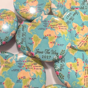 Puede incluir: Una colección de chapas redondas con un diseño de mapa mundial, océano azul claro y continentes de color tostado. Cada chapa muestra pequeños iconos de corazones rosas marcando varias ubicaciones y el texto Jesus-The Way 2027.