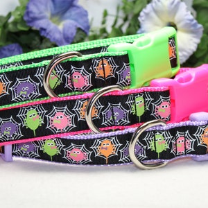 May include: Three dog collars with a black background and a spiderweb pattern. The collars are in different colors: green, pink, and purple. Each collar has a silver ring and a plastic buckle.