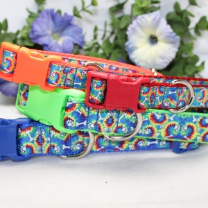 May include: A stack of four dog collars with colorful tie-dye patterns and paw prints. Each collar has a different colored plastic buckle: orange, red, green, and blue. The collars are made of a durable fabric.