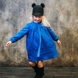 May include: A young girl wearing a blue dress with a black hat with fuzzy ear flaps. She is standing on a wooden floor in front of a textured wall.