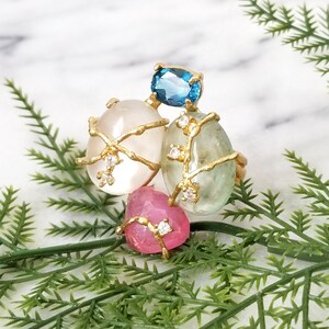 Peut inclure: Une bague en or avec un groupe de pierres précieuses de forme ovale. La bague présente une pierre blanche, vert clair et rose, chacune sertie dans une cage de fil d'or. Les pierres sont rehaussées de petits diamants clairs.
