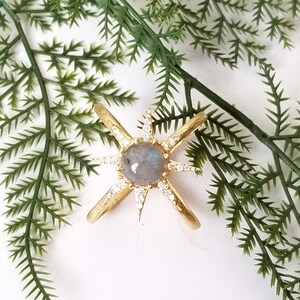 Peut inclure: Bague en or avec une grande pierre de labradorite grise au centre. La bague est en forme d'étoile avec de petites pierres claires autour de la labradorite.