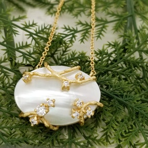 May include: A gold necklace with a white oval pendant featuring a delicate branch design with small clear crystals.