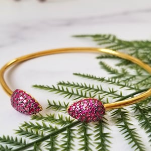 May include: A gold bangle bracelet with two pink gemstone accents. The bracelet is open and has a simple design.