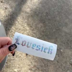 Puede incluir: Una pegatina blanca con la palabra "Lovesick" en una fuente holográfica iridiscente. La pegatina es rectangular y está sujeta por una mano con esmalte de uñas negro.