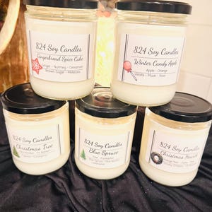 May include: Five glass jars of soy candles with black lids are stacked. Each jar has a label with the text "824 Soy Candles" and a scent name: Gingerbread Spice Cake, Winter Candy Apple, Christmas Tree, Blue Spruce, and Christmas Hearth.