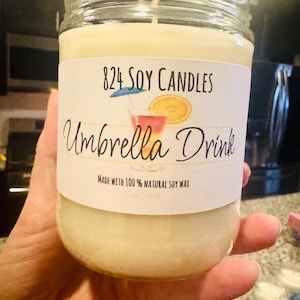 May include: A jar candle with a cream-colored wax and a label that reads "824 Soy Candles Umbrella Drink." The label features a cocktail illustration with an orange slice and a small blue umbrella. The text "Made with 100% Natural Soy Wax" is also visible.