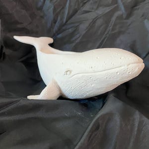 May include: A white ceramic whale figurine with a textured surface. The whale has a detailed eye and fin, and a small opening on its back. The figurine is resting on a black fabric surface.