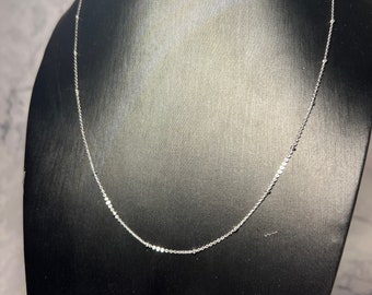 May include: A delicate silver necklace with small, evenly spaced, round embellishments. The necklace is displayed on a black mannequin, showcasing its design and length. The background is a light gray and white marble pattern.