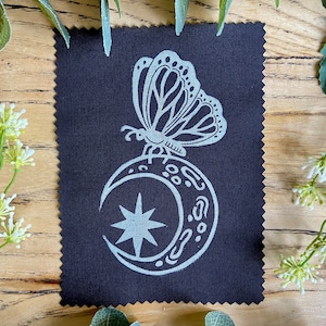 May include: A dark blue fabric square featuring a light grey butterfly design atop a crescent moon and star. The fabric edges are scalloped. Decorative greenery surrounds the fabric.