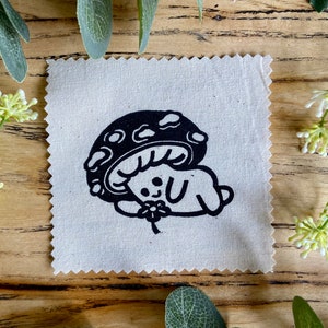 May include: A black and white illustration of a bunny wearing a mushroom hat and holding a flower. The illustration is printed on a beige fabric square.
