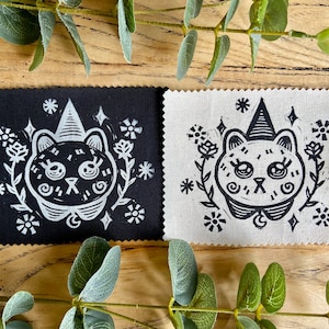 May include: Two black and white fabric squares with a hand-printed design of a cat wearing a witch's hat and surrounded by flowers. The cat is white on a black background on one square and black on a white background on the other square.