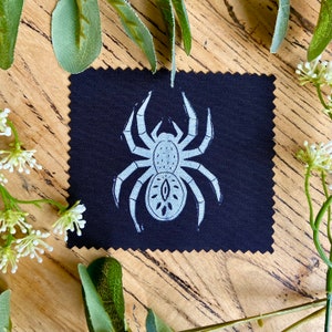 May include: A black fabric coaster featuring a white spider design. The spider has a detailed design with a large eye in the centre of its body.
