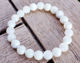 May include: A white beaded bracelet with round, polished beads. The bracelet is resting on a wooden surface, with a blurred green background. The beads appear to be made of a pearlescent material.
