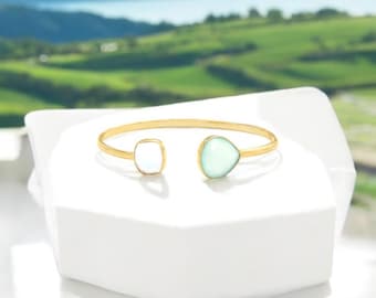 May include: A gold bangle bracelet with a light blue teardrop-shaped gemstone and a white square gemstone.