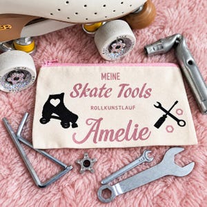 Op de afbeelding: Een beige canvas etui met rits en roze rand, voorzien van de tekst "MEINE Skate Tools", "ROLLKUNSTLAUF" en "Amelie" in roze en zwart. Het ontwerp bevat een zwart silhouet van een rolschaats met een hartje en een afbeelding van gereedschap.
