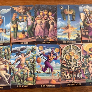Puede incluir: Diez cartas de tarot con ilustraciones de fantasía. Las cartas muestran figuras, símbolos y texto como "Ace of Cups", "2 of Cups" y "The Lovers". La obra de arte utiliza azules, morados y naranjas. Las cartas están dispuestas sobre una superficie de madera.