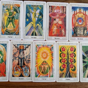 May include: A collection of ten tarot cards, each with unique illustrations and titles. The cards feature vibrant colors and symbolic imagery, including figures, celestial bodies, and natural elements. The cards are arranged on a wooden surface.