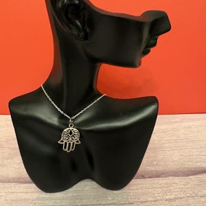 May include: A silver necklace featuring a Hamsa hand pendant. The pendant has an openwork design and is suspended from a fine chain. The necklace is displayed on a black mannequin against a red backdrop.