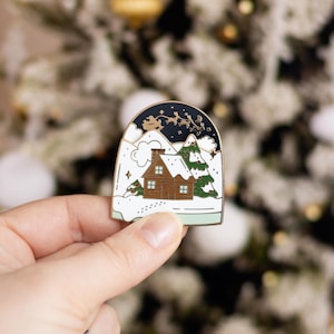 May include: A Christmas-themed enamel pin featuring a snow globe design. The pin depicts a cozy brown cabin with snow-covered roof and evergreen trees. The top of the pin shows a dark blue night sky with a silhouette of Santa's sleigh and reindeer.