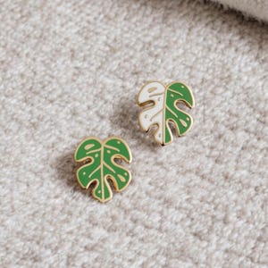 May include: Two gold-tone enamel pins shaped like monstera leaves. One leaf is green with white spots, and the other is white with green spots.