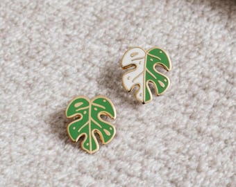 May include: Two gold-tone enamel pins shaped like monstera leaves. One leaf is green with white spots, and the other is white with green spots.